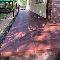 Wykończony taras z brązowych płytek ceramicznych, widok z boku, z elementami metalowej konstrukcji i fragmentem elewacji budynku wykończonej cegłą.