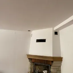 Białe ściany i sufit po malowaniu, fragment kominka z drewnianym blatem i kamienną obudową, widoczne otwory wentylacyjne w ścianie i suficie.