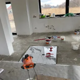 Układanie płytek marmurowych na podłodze w surowym wnętrzu, widoczna mieszarka do kleju, przyssawki do płytek oraz narzędzia glazurnicze.