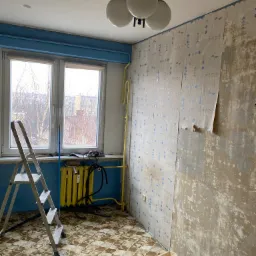 Pok&oacute;j w trakcie usuwania starej tapety, widoczne fragmenty oderwanej tapety na ścianie, drabina oparta o okno, grzejnik pomalowany na ż&oacute;łto i wiadro z wodą na podłodze.