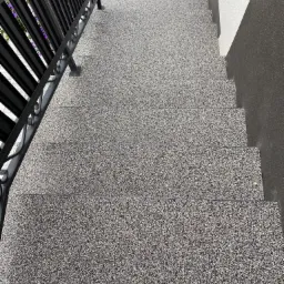 Schody zewnętrzne pokryte szarą, kamyczkową żywicą epoksydową, z czarną, metalową balustradą i fragmentem elewacji w odcieniach szarości.