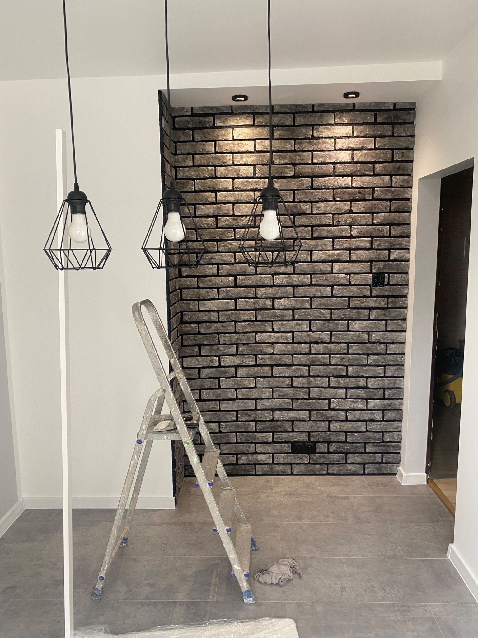 Ściana wykończona szarą cegłą dekoracyjną, widoczne trzy lampy wiszące w stylu industrialnym i drabina aluminiowa, szara podłoga z płytek.