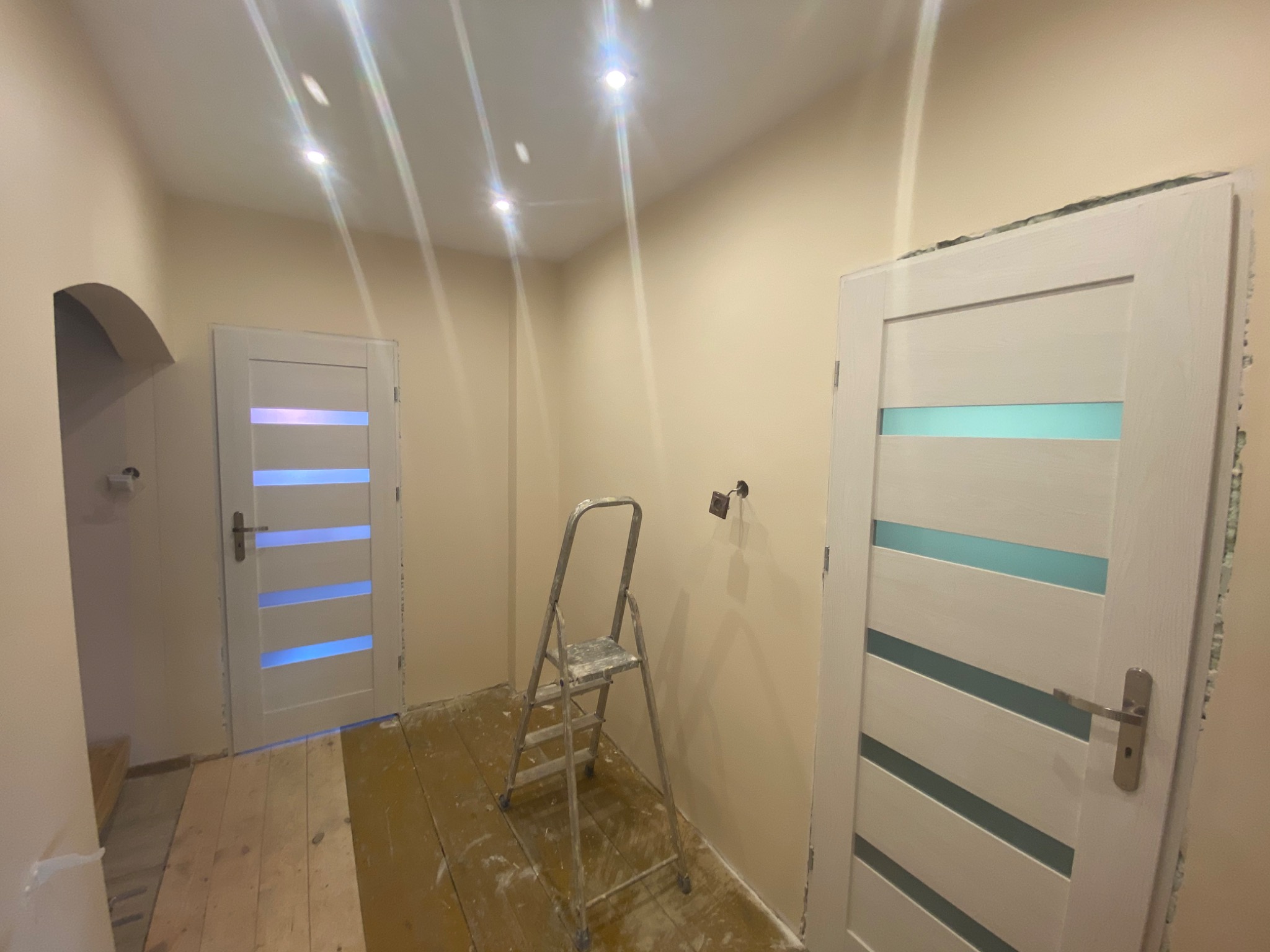 Wnętrze w trakcie remontu: pomalowane ściany na beżowo, dwie białe drzwi z poziomymi przeszkleniami, aluminiowa drabina stojąca na zabezpieczonej podłodze, widoczne puszki elektryczne.