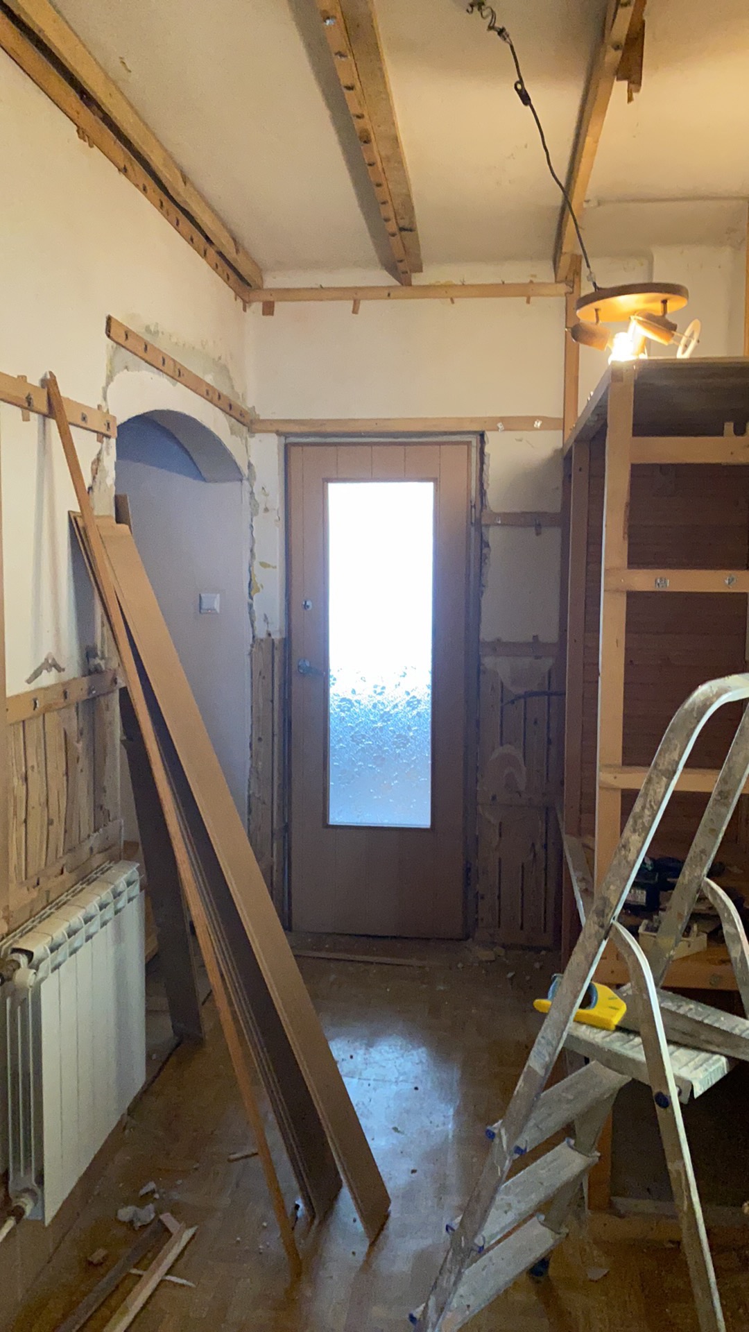 Wnętrze w trakcie demontażu boazerii i przygotowania do remontu, widoczne odsłonięte fragmenty ścian, grzejnik, drzwi z szybą ornamentową, listwy przypodłogowe oparte o ścianę oraz drabina malarska...