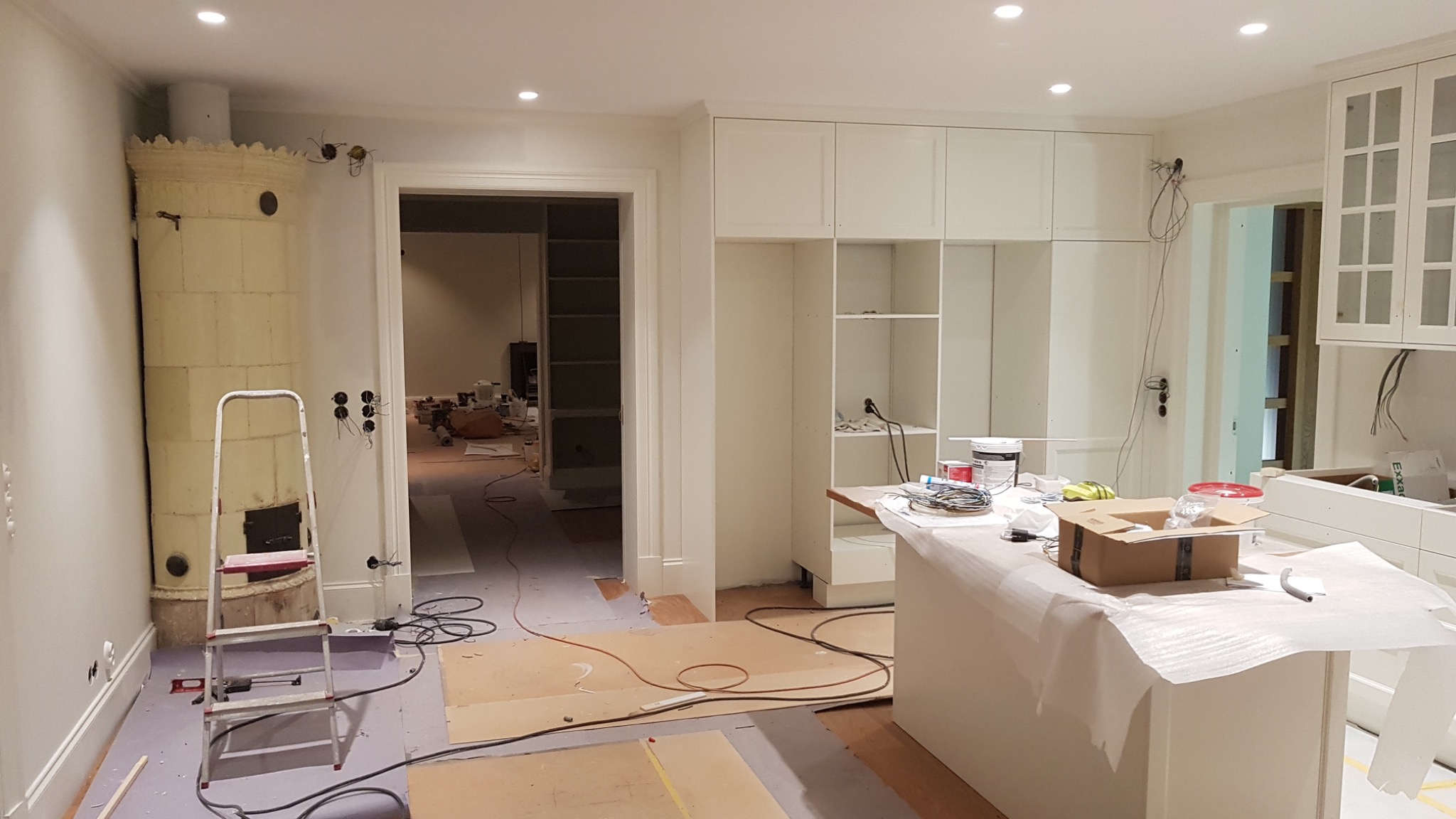Wnętrze w trakcie prac wykończeniowych: widoczny piec kaflowy, drabina, podłoga zabezpieczona kartonem, częściowo zamontowane białe szafki kuchenne i wyspa, kable wystające ze ściany, otwarte...