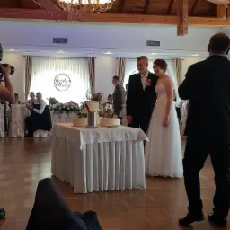 Para młoda w eleganckich strojach pozuje obok tortu weselnego na sali bankietowej z drewnianym sufitem i żyrandolem, fotograf uwiecznia moment.