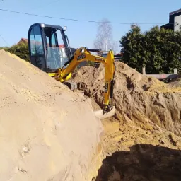 Żółta mini koparka JCB na skraju wykopu w piaszczystej ziemi, widoczne domy w tle, słoneczny dzień.