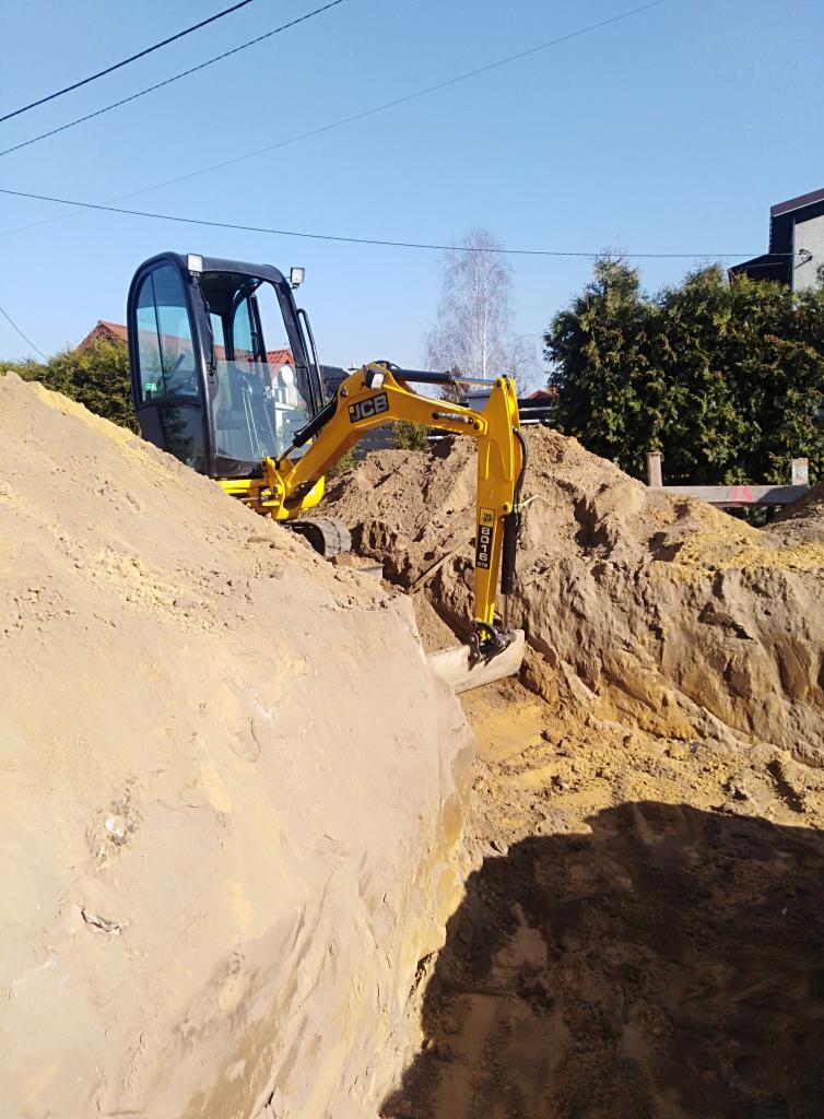 Żółta mini koparka JCB na skraju wykopu w piaszczystej ziemi, widoczne domy w tle, słoneczny dzień.