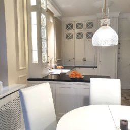 Marzena Camila Wojtkiewicz GREEN-ARTE pracownia wnętrz i malarstwa - Jasna kuchnia w klasycznym stylu z czarnym blatem, owocami i dekoracyjną lampą. Białe krzesła przy okrągłym stole. Subtelne detale wykończenia.