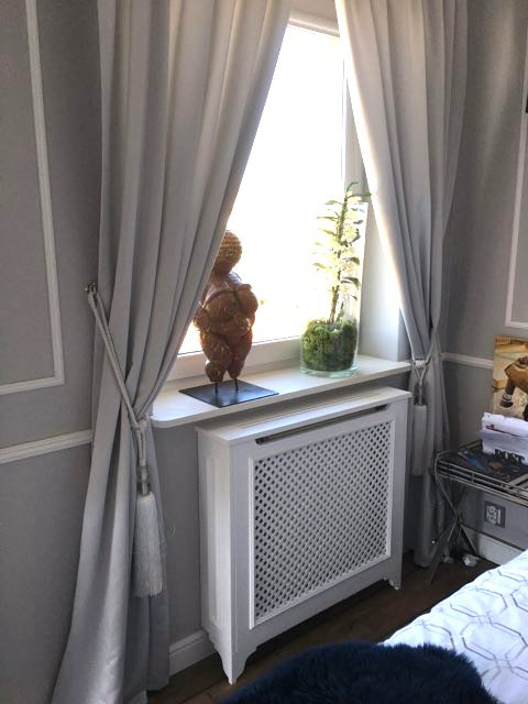 Jasne zasłony z dekoracyjnym wiązaniem, rzeźba i roślina na parapecie nad zabudowanym grzejnikiem w pokoju o szarych ścianach z dekoracyjnymi listwami.