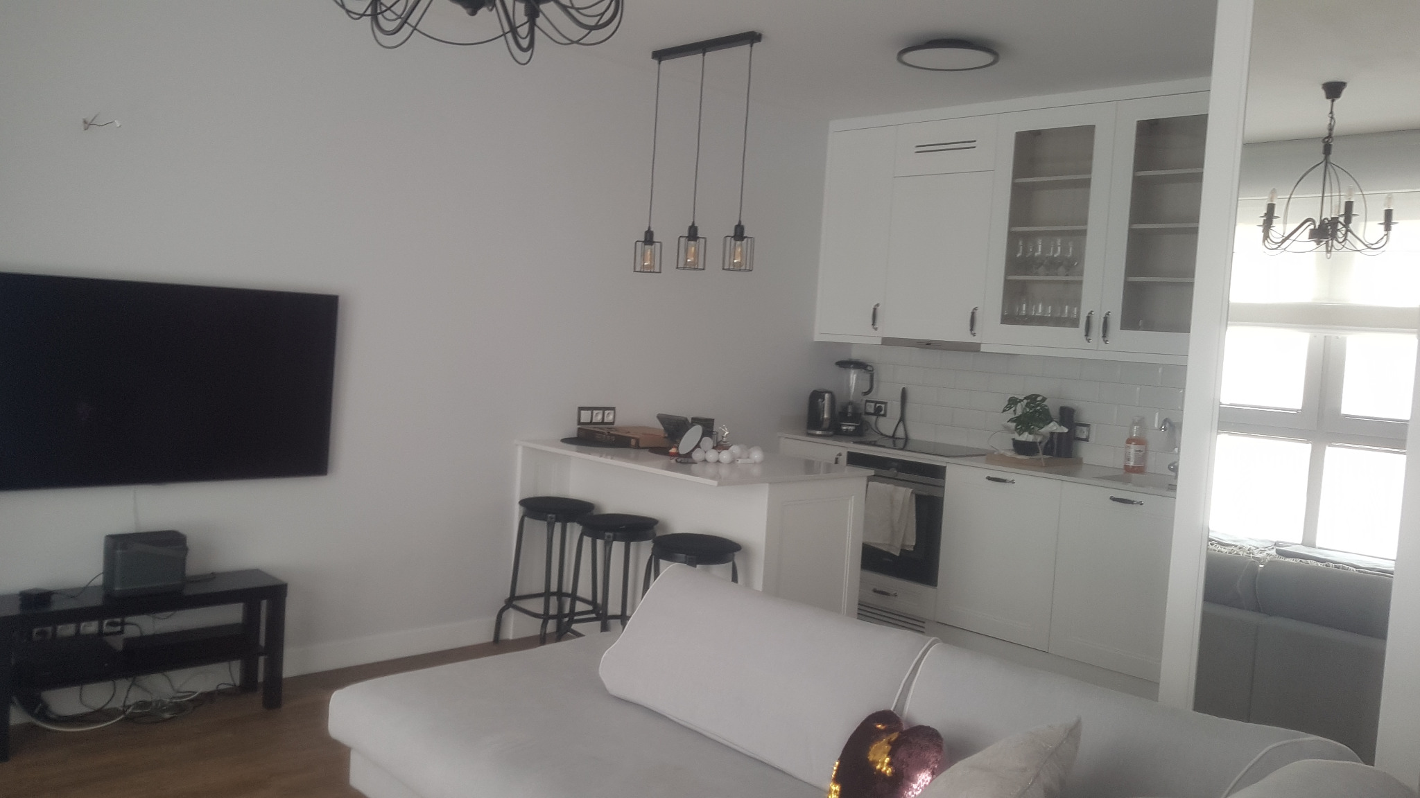 Jasne wnętrze: salon z aneksem kuchennym w bieli. Minimalistyczne lampy wiszące nad barkiem, telewizor na ścianie, sofa z poduszkami. Nowoczesny design.
