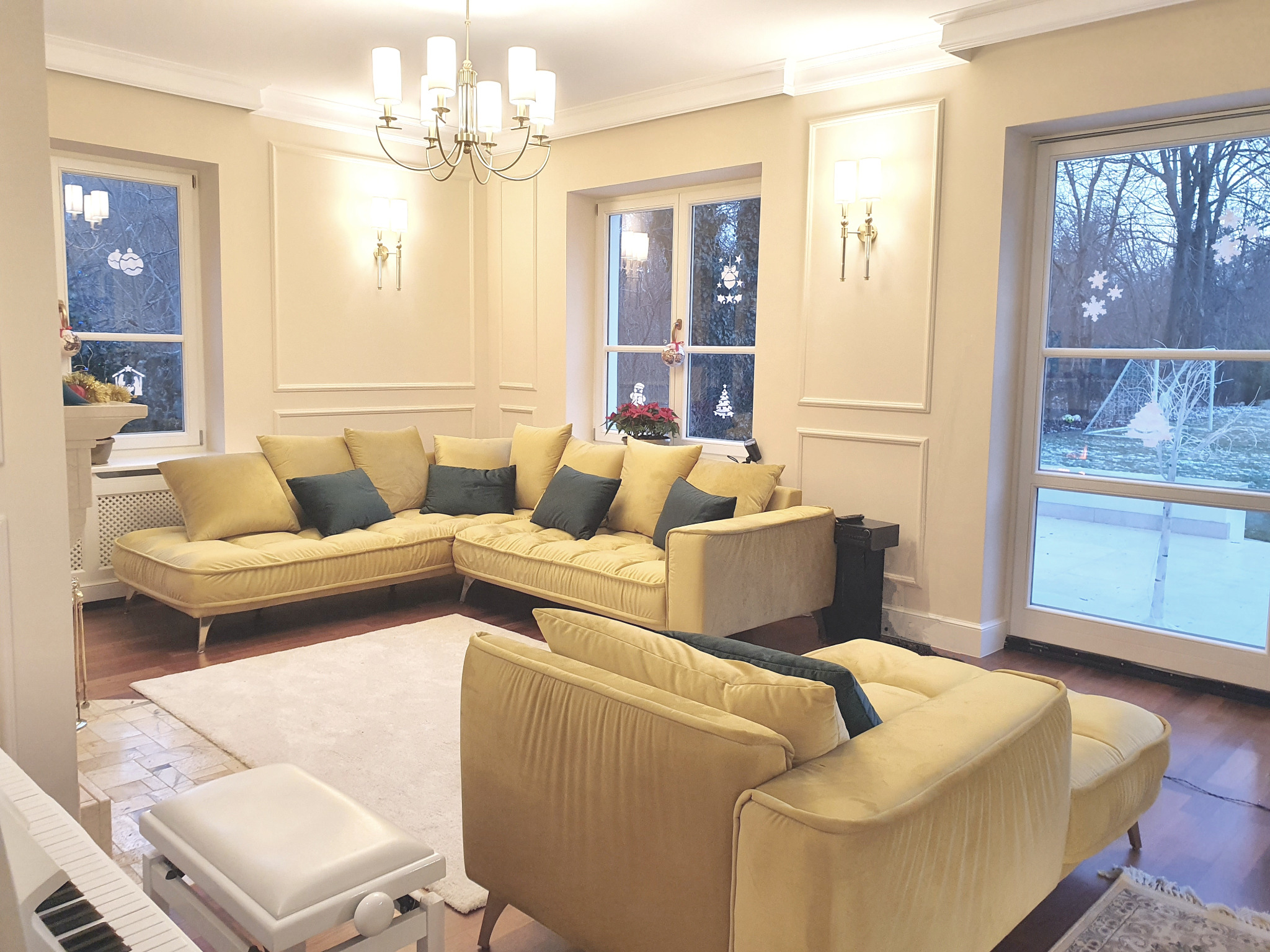Elegancki salon z żółtą sofą narożną, białym dywanem i pianinem. Świąteczny akcent: dekoracje okienne. Ciepłe oświetlenie podkreśla klasyczny wystrój.