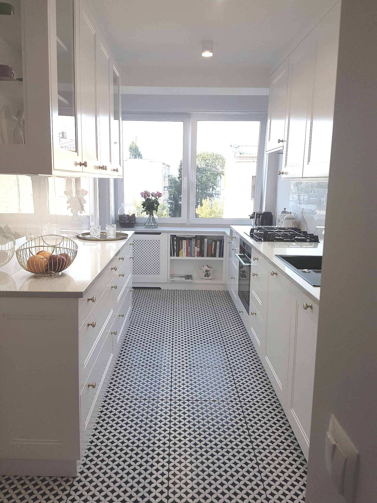 Wąska, jasna kuchnia z białymi szafkami i wzorzystą podłogą. Na blacie kosz z owocami, w tle okno z widokiem na zieleń. Minimalistyczny styl.