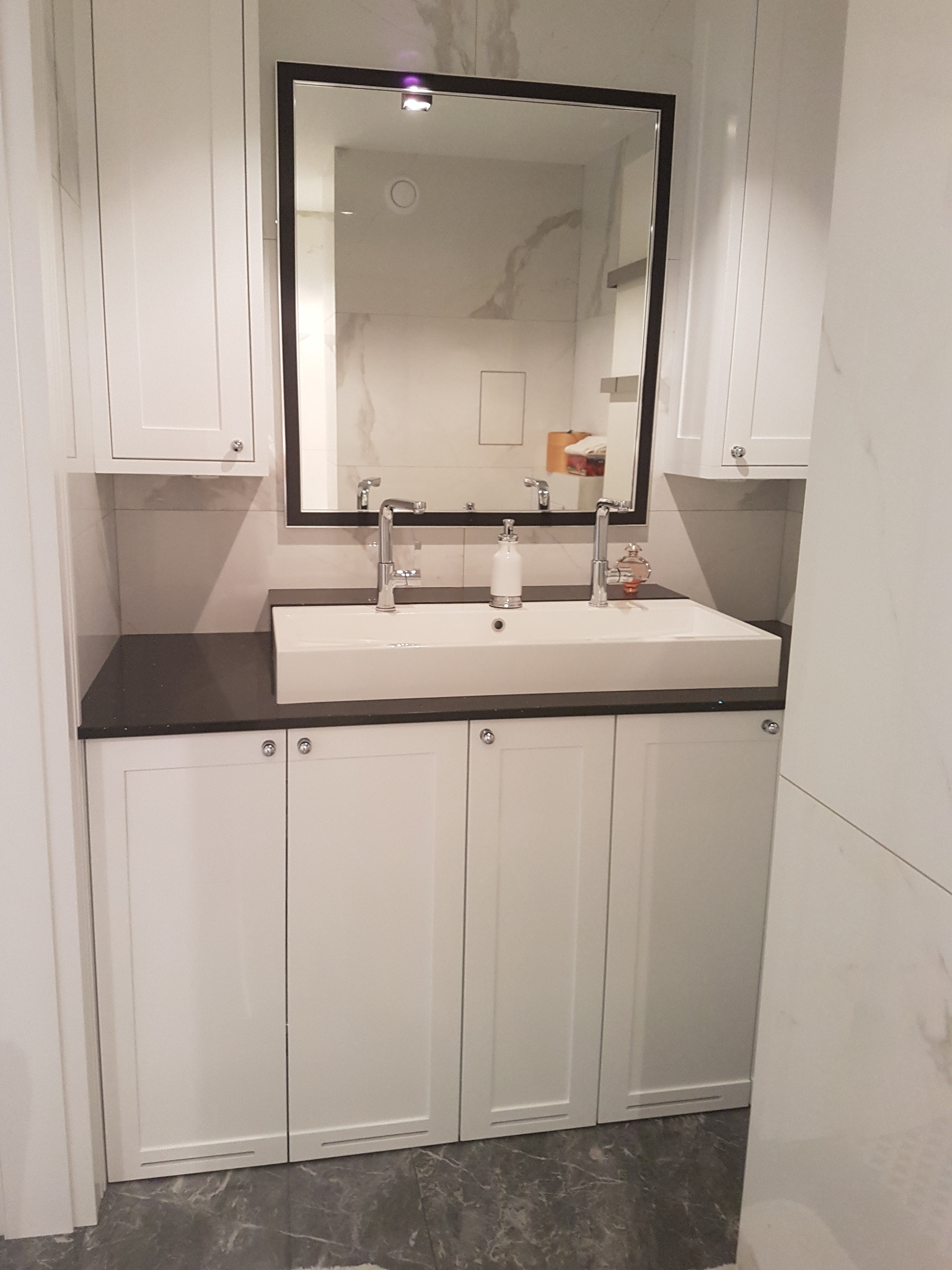 Nowoczesna łazienka z białą szafką pod umywalką, czarnym blatem i dużym lustrem w czarnej ramie. Widoczne baterie umywalkowe i marmurowe płytki na ścianach.