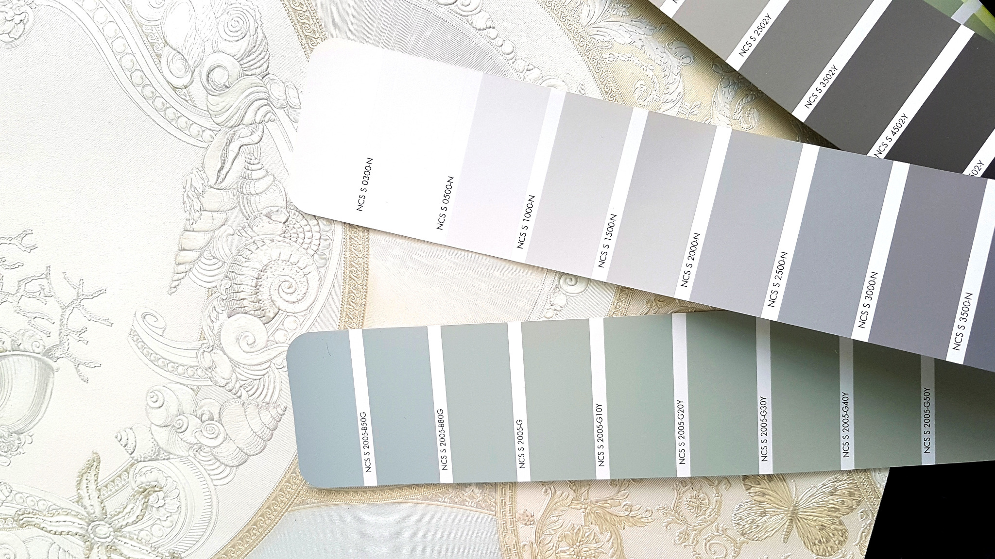 Paleta kolorów NCS na tle tapety z ornamentami. Odcienie szarości i zieleni. Wybór barw do aranżacji wnętrz. Detal dekoracyjny, wzornik kolorystyczny.