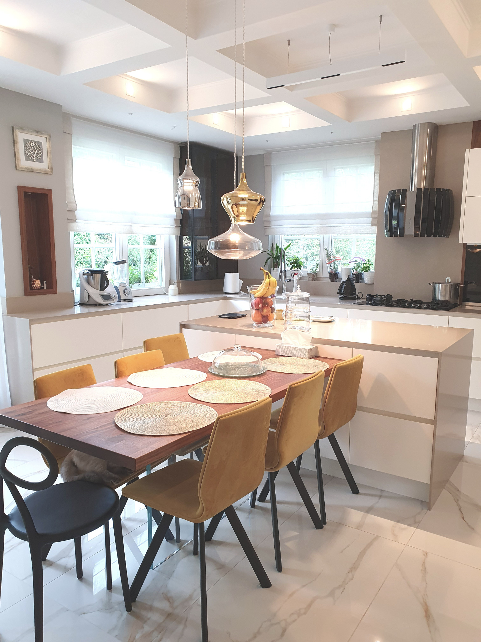 Jasna kuchnia z wyspą i stołem jadalnym z żółtymi krzesłami. Nad stołem wiszą designerskie lampy. Marmurowa podłoga i okno z roletami rzymskimi.