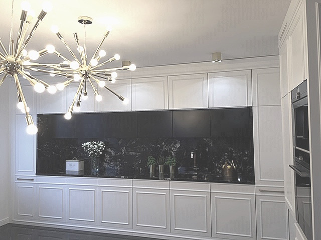 Nowoczesna kuchnia z białymi szafkami w stylu klasycznym, czarnym blatem i marmurowym panelem. Nad blatem dekoracyjna lampa w stylu glamour.