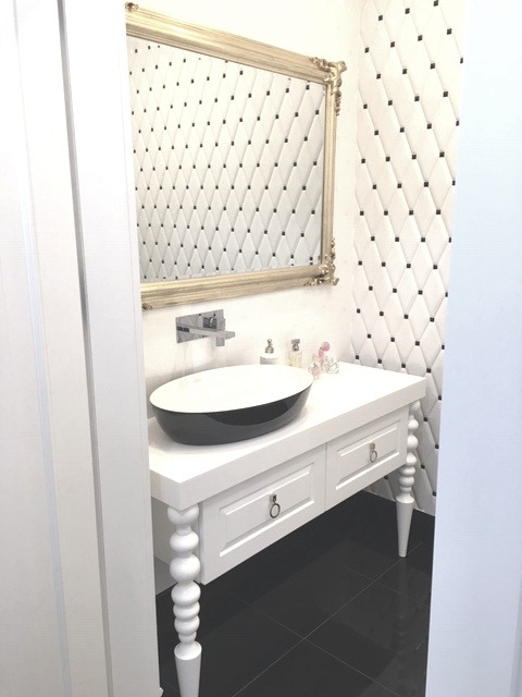 Elegancka łazienka z czarno-białą armaturą, owalną umywalką na białej konsoli z frezowanymi nogami i lustrem w złotej ramie na tle ściany w romby.