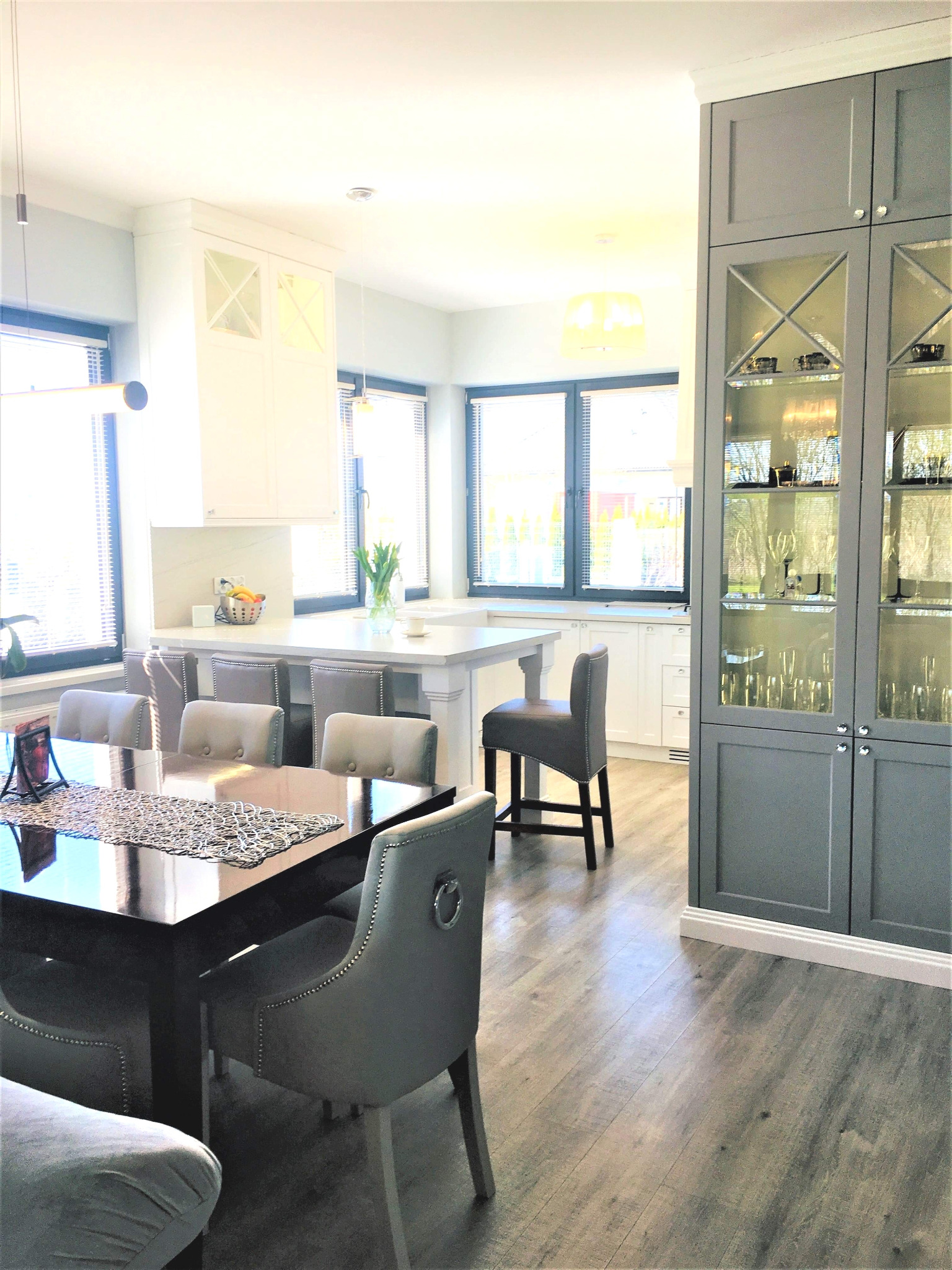 Jasna kuchnia z wyspą i szarymi krzesłami, widok na przeszkloną szafę z zastawą. Nowoczesne oświetlenie i drewniana podłoga dodają elegancji.