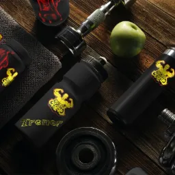 Zestaw gadżetów dla trenera personalnego: butelki, ręcznik, pojemnik na odżywki z logo siłowni, hantle i zielone jabłko na drewnianym stole.