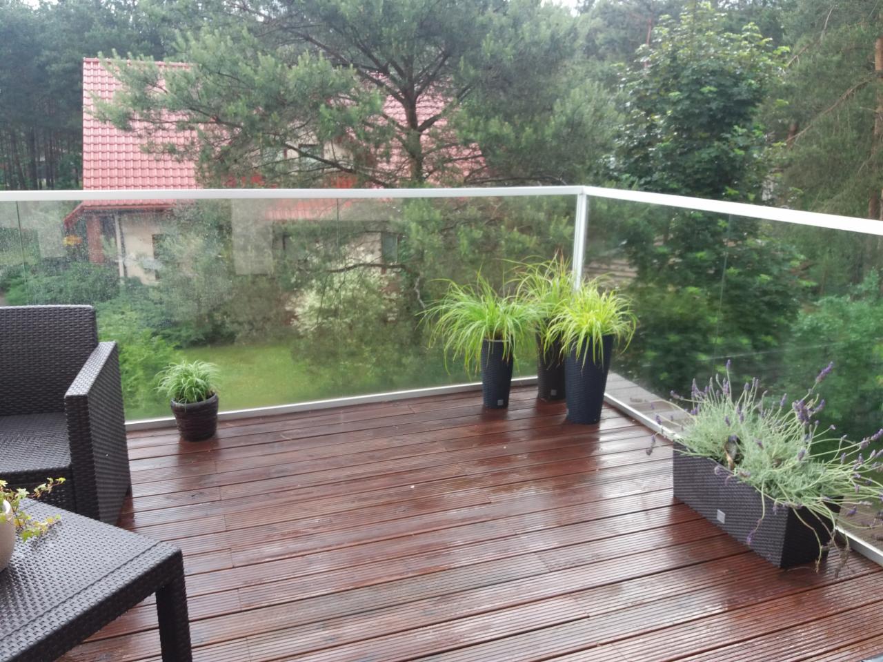 Balkon z drewnianą podłogą i szklaną balustradą, widok na ogród i dom z czerwonym dachem w tle, rośliny w donicach na balkonie.