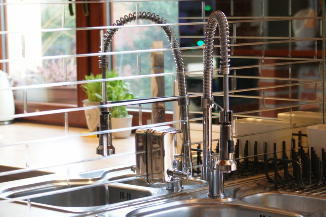 Błyszcząca, nowoczesna bateria kuchenna z wyciąganą wylewką, widziana przez siatkę metalowych półek suszarki do naczyń, z oknem w tle.