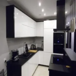 Nowoczesna kuchnia w czarno-białej kolorystyce z błyszczącymi frontami szafek, czarnym blatem i sprzętami AGD, minimalistyczny okap, oświetlenie punktowe.