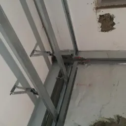 Metalowy stelaż sufitowy przymocowany do nierównego, starego sufitu z widocznymi instalacjami i ubytkami tynku.