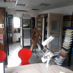 Ekspozycja w salonie: próbki kolorów, ramy okienne, drzwi wewnętrzne z przeszkleniami, rolety wewnętrzne Hanarol, butelka płynu do czyszczenia Clin na stole.