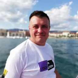 Mężczyzna w białej koszulce z logo 'COALITION' na tle morza, błękitnego nieba i zabudowań miasta. Uśmiechnięta twarz, naturalne światło.