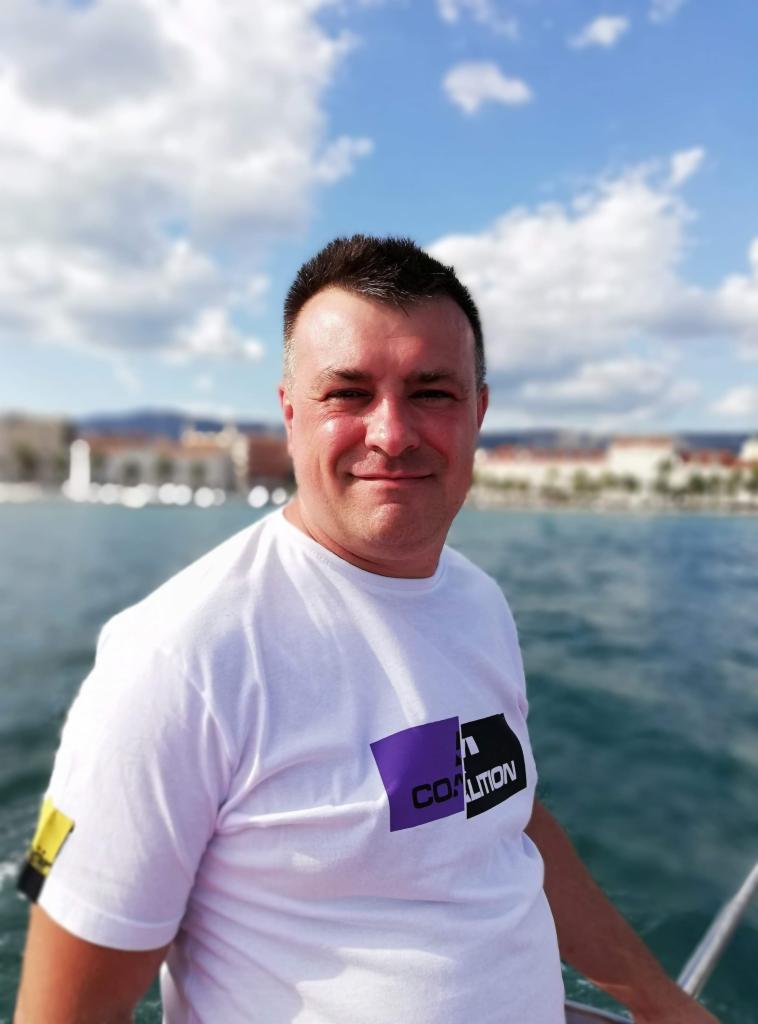 Mężczyzna w białej koszulce z logo 'COALITION' na tle morza, błękitnego nieba i zabudowań miasta. Uśmiechnięta twarz, naturalne światło.
