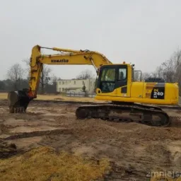 Żółta koparka Komatsu PC240LC na placu budowy z wykopami w ziemi, budynek w tle, pochmurne niebo.