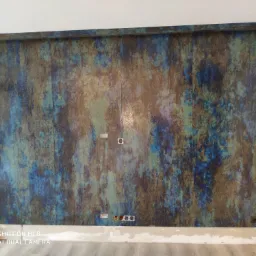 Ściana wyłożona panelami imitującymi rdzewiejący metal w odcieniach niebieskiego, brązowego i zielonego, z widocznymi puszkami elektrycznymi i gniazdkami, na podłodze jasne panele.