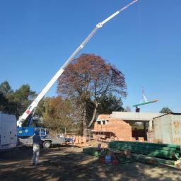 PHU TEGOLA Bartosz Wąsik - Budowa domu w trakcie wznoszenia, dźwig podnosi element konstrukcyjny, widoczne cegły, drewniane belki i pracownik na dachu.
