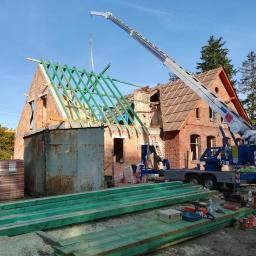 PHU TEGOLA Bartosz Wąsik - Ceglany dom w trakcie remontu dachu, z widoczną konstrukcją więźby dachowej, dźwigiem i stertą przygotowanych belek na pierwszym planie.