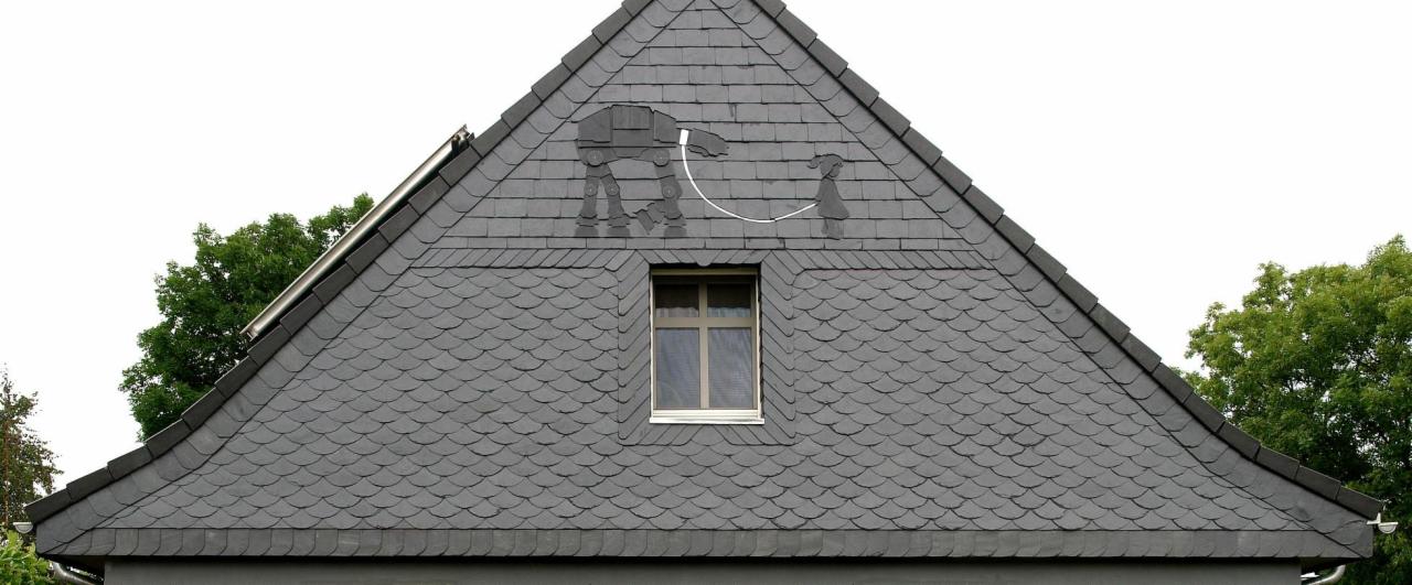 Szary dach pokryty łupkiem z dekoracyjnym motywem: AT-AT ze Star Wars prowadzący dziewczynkę na smyczy, umieszczony nad oknem mansardowym.