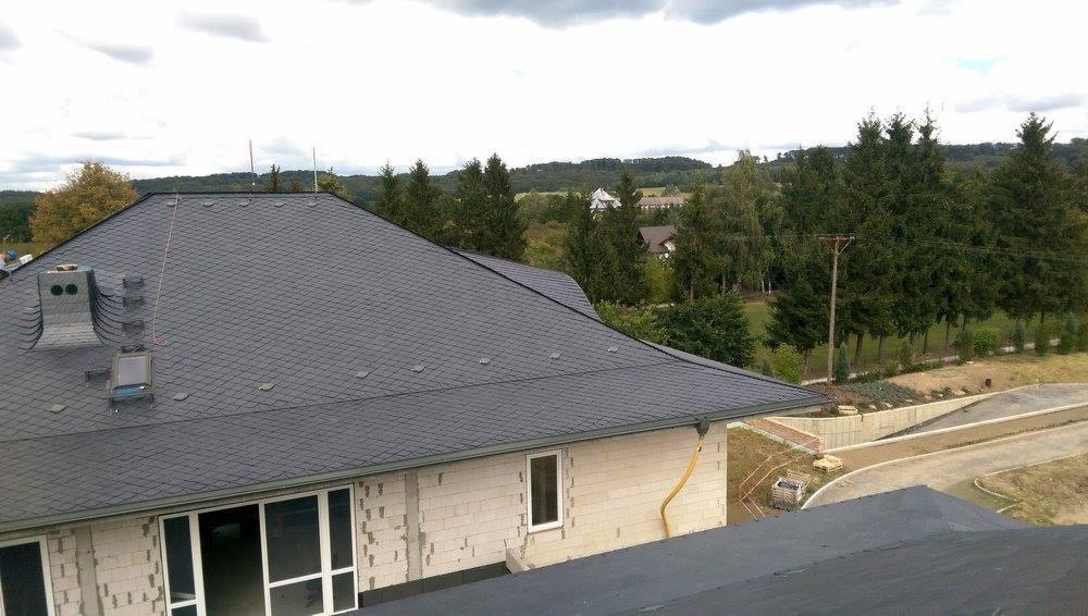 Nowy, ciemnoszary dach z łupka na domu jednorodzinnym w stanie surowym, widok z góry na komin, okna i rynny, w tle zieleń drzew i pochmurne niebo.