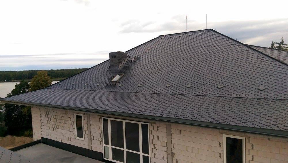 Szary dach z łupka naturalnego na domu w trakcie budowy, widoczny komin, okno dachowe i rynna, w tle jezioro i las.