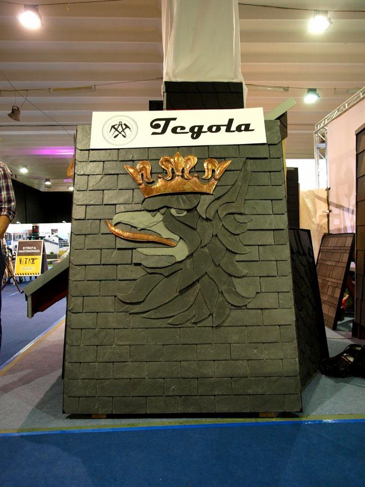 Ekspozycja firmy Tegola prezentująca wzór dachu z motywem orła i korony, wykonany z ciemnych płytek dachowych na stoisku targowym.