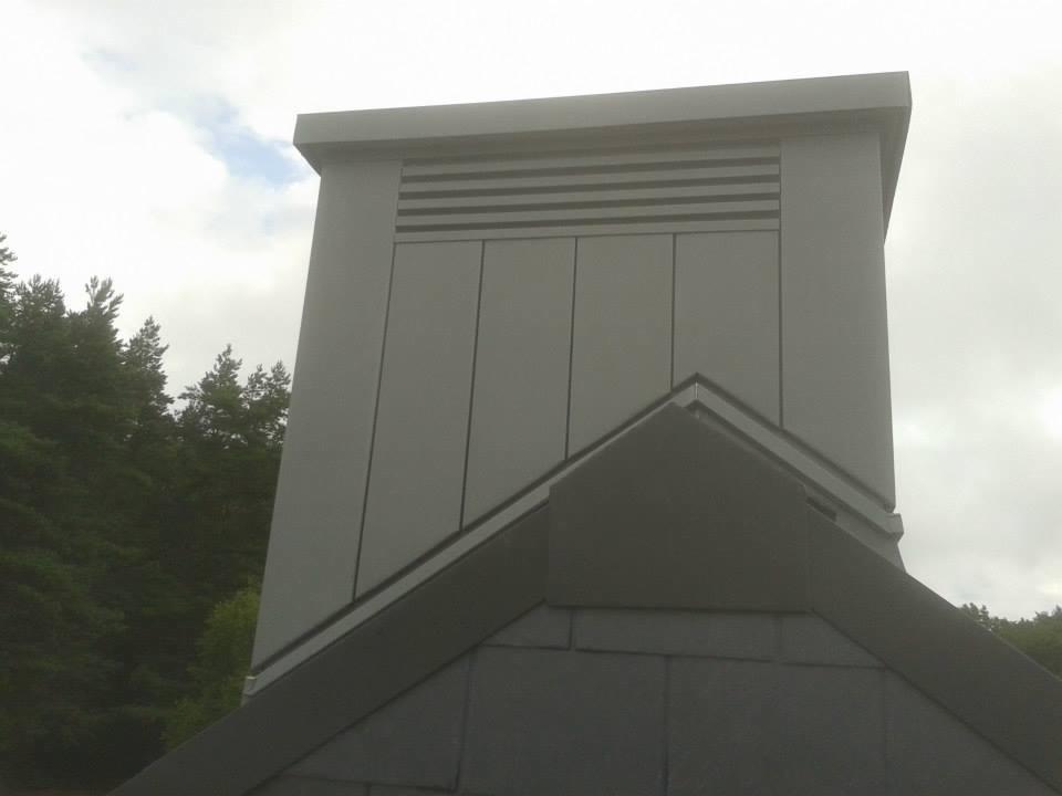 Szary komin wentylacyjny z metalową obudową, widoczny z perspektywy dachu pokrytego ciemnoszarą dachówką, na tle pochmurnego nieba i zieleni drzew.