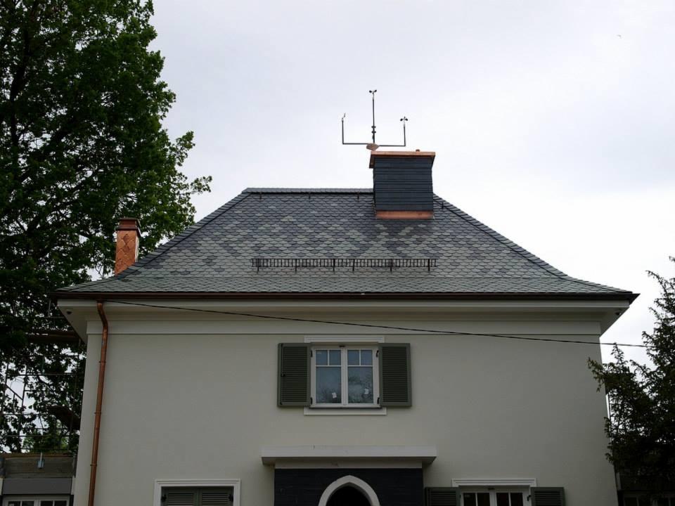 Dom z dachem pokrytym łuską w odcieniach szarości, z miedzianymi rynnami i kominem z elementami miedzianymi, zakończonym instalacją odgromową.