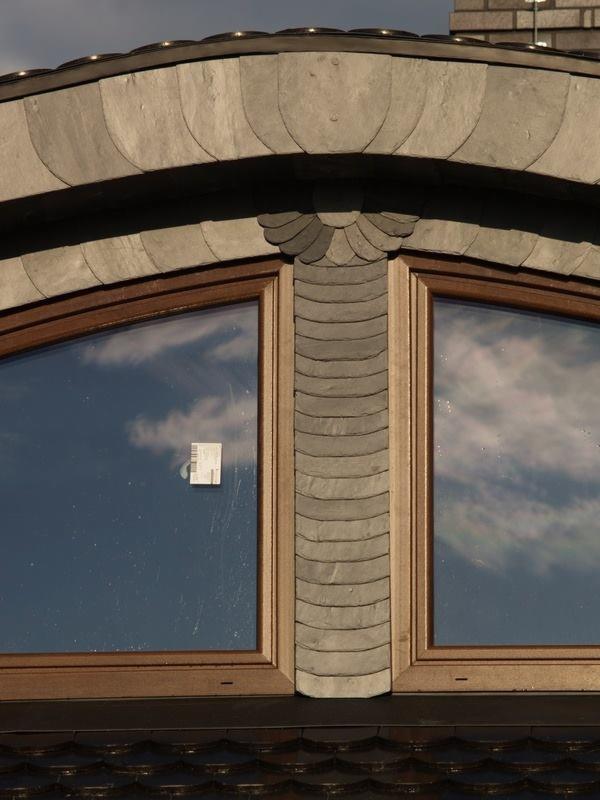Fragment dachu z lukarną pokrytą łupkiem, z widocznym odbiciem chmur w oknach i naklejką na szybie.