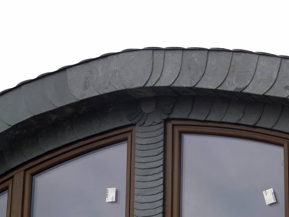Fragment okna z łukowatym nadprożem pokrytym dekoracyjnym, łupkowym pokryciem dachowym w kształcie rybiej łuski. Widoczne brązowe ramy okienne i szare szyby z naklejkami.