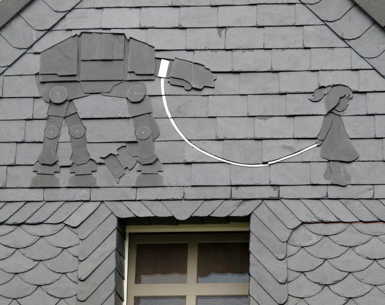 Dach kryty łupkiem z dekoracyjnym motywem: postać AT-AT z Gwiezdnych Wojen połączona białą linią z sylwetką dziewczynki skaczącej na skakance, widok na fragment okna dachowego.