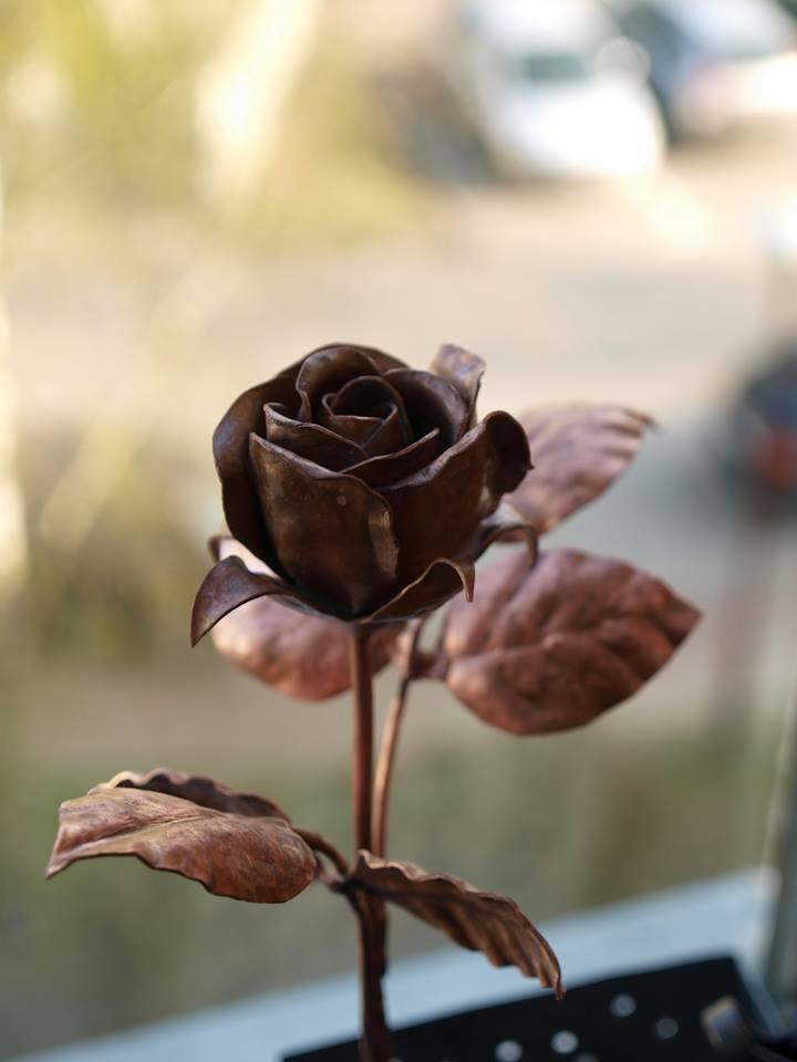 Ręcznie wykonana, miedziana róża z detalami blachy, stojąca na parapecie okna z rozmytym widokiem na zewnątrz.