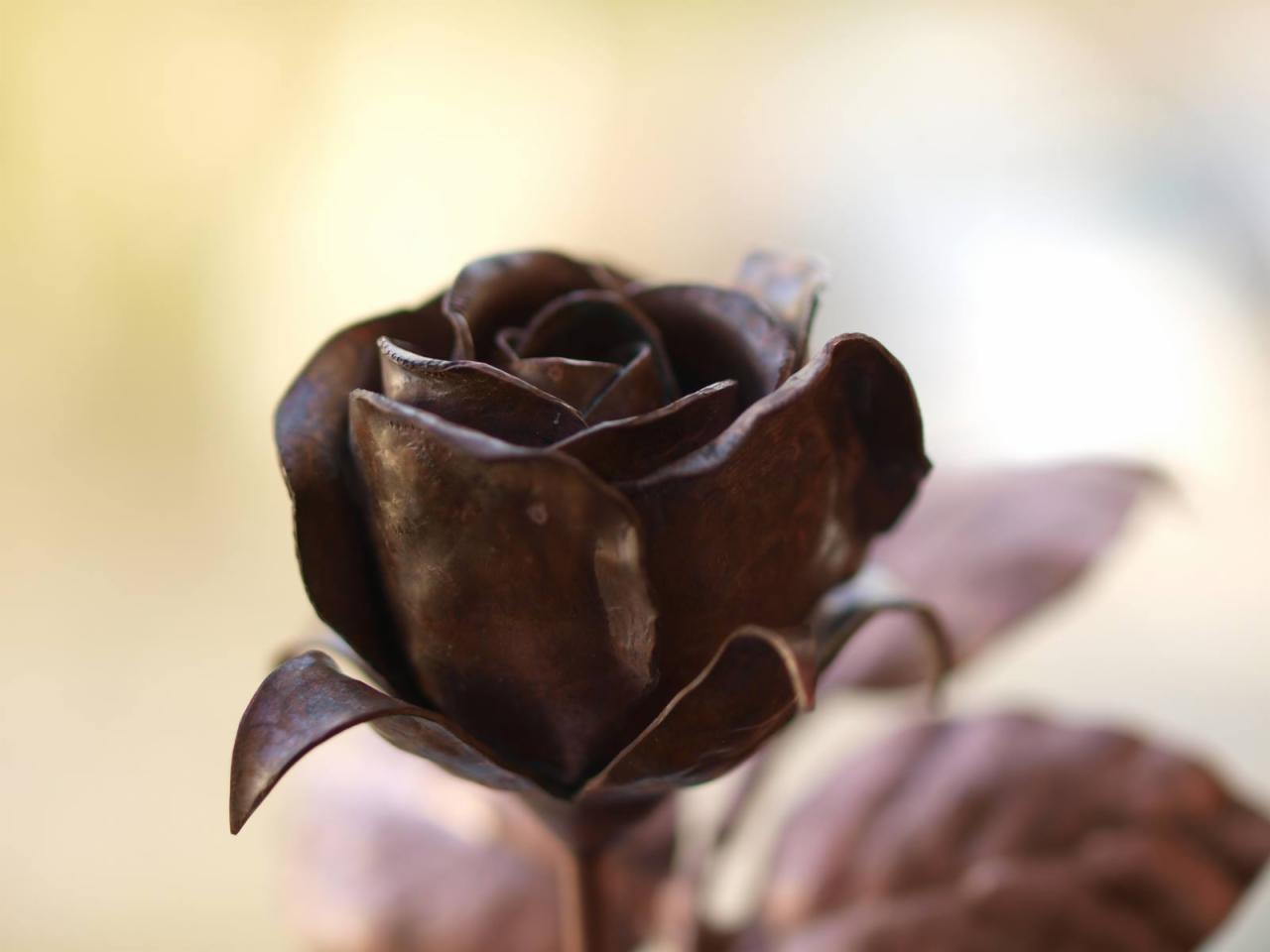 Dekoracyjna róża z metalu o ciemnobrązowym odcieniu, z widocznymi detalami płatków i liści, na rozmytym, jasnym tle.