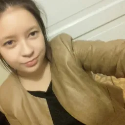 Portret młodej kobiety w brązowej kurtce na tle białych drzwi i wzorzystej pościeli.