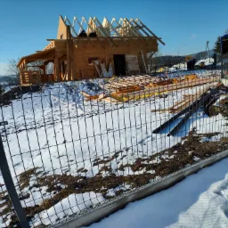 Surowa konstrukcja drewnianego domu w trakcie budowy, widoczna przez metalowe ogrodzenie, z elementami konstrukcyjnymi dachu wznoszącymi się ku błękitnemu niebu, otoczona śnieżnym krajobrazem.