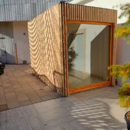 Nowoczesna, drewniana konstrukcja z dużym oknem, prawdopodobnie sauna, ustawiona na tarasie z płytek w słoneczny dzień. Widoczny fragment zadaszenia i iglaste drzewko w donicy.