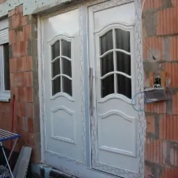 Białe, dwuskrzydłowe drzwi wejściowe z przeszkleniami w trakcie montażu w Białymstoku, widoczne niedokończone ściany z cegły i okno z częściowo opuszczoną roletą.